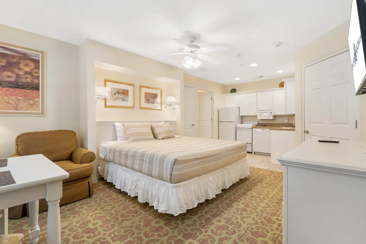 This image shows a king bedroom suite featuring a comfortable bed, an armchair, a kitchenette, and tasteful wall art. Natural light filters through the windows, creating a cozy and inviting atmosphere. The kitchenette area is conveniently positioned within the suite, adding versatility for guests who want snacks or beverages close at hand.