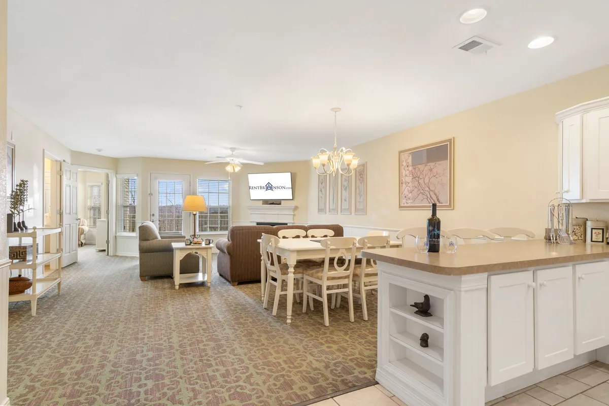 This image captures an inviting interior open-plan area featuring a dining table, a plush sofa, a kitchen island, and a glowing chandelier. The space is softly lit, exuding warmth and togetherness, ideal for family meals, entertaining guests, or relaxing evenings. The combination of seating and dining spaces exemplifies the heart of the home where people naturally gather.