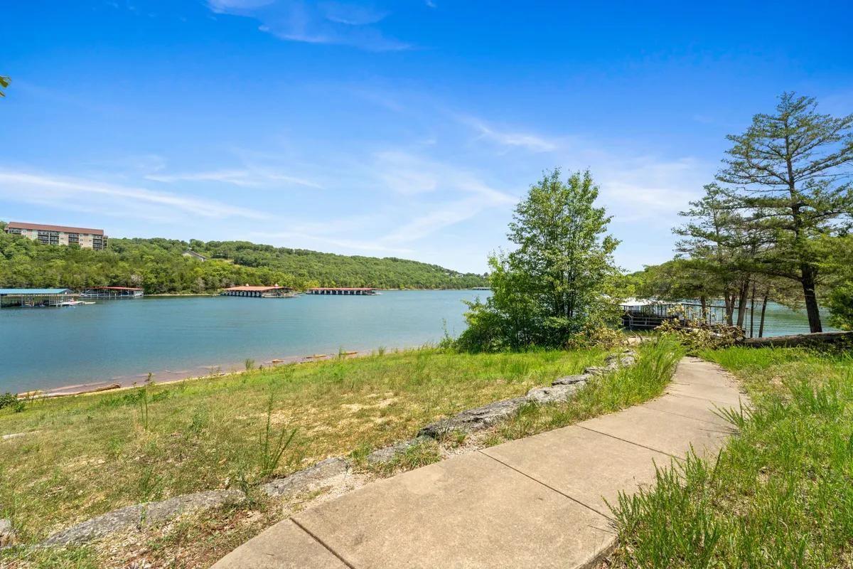 A picturesque view of calm lake waters surrounded by vibrant greenery and distant rolling hills. A pathway winds along the shoreline, offering spots for fishing, picnicking, or simply enjoying nature. The serene mood captures the inviting atmosphere of Table Rock Lake.