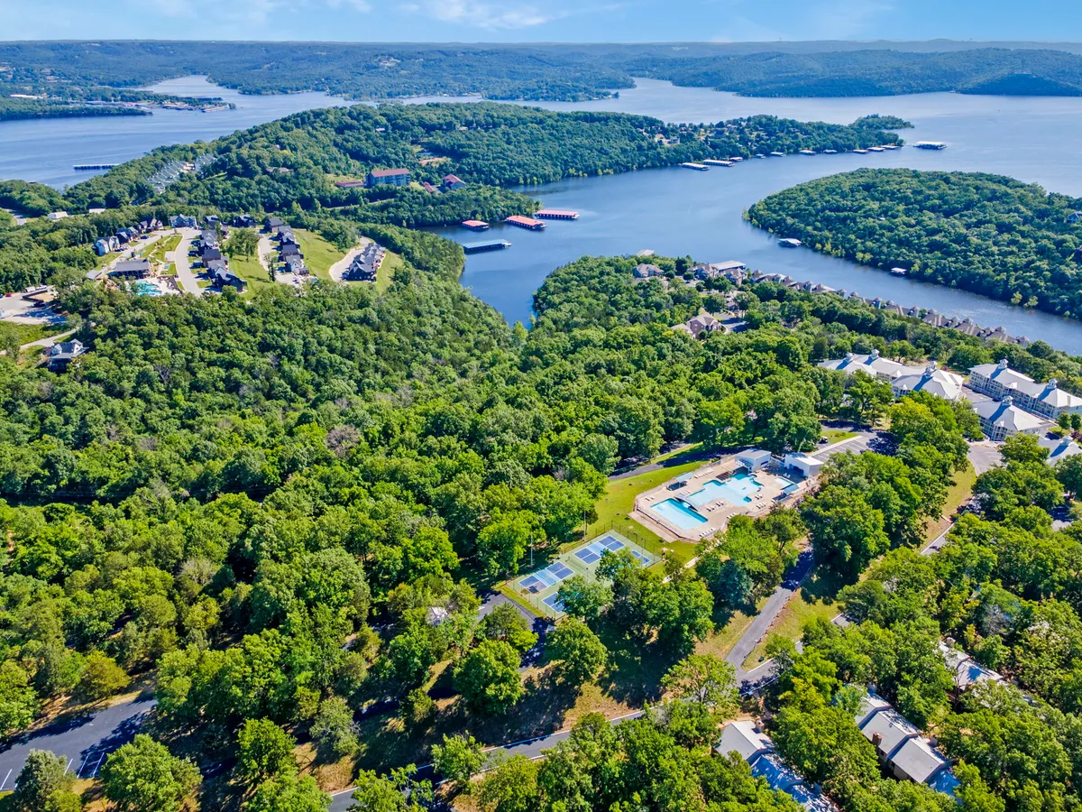 A panoramic view showcases the resort’s swimming pools, tennis courts, wooded areas, and a scenic lake with residential buildings nestled among the trees. The composition highlights multiple amenities within a single expansive landscape, illustrating the resort’s vibrant community and natural beauty.