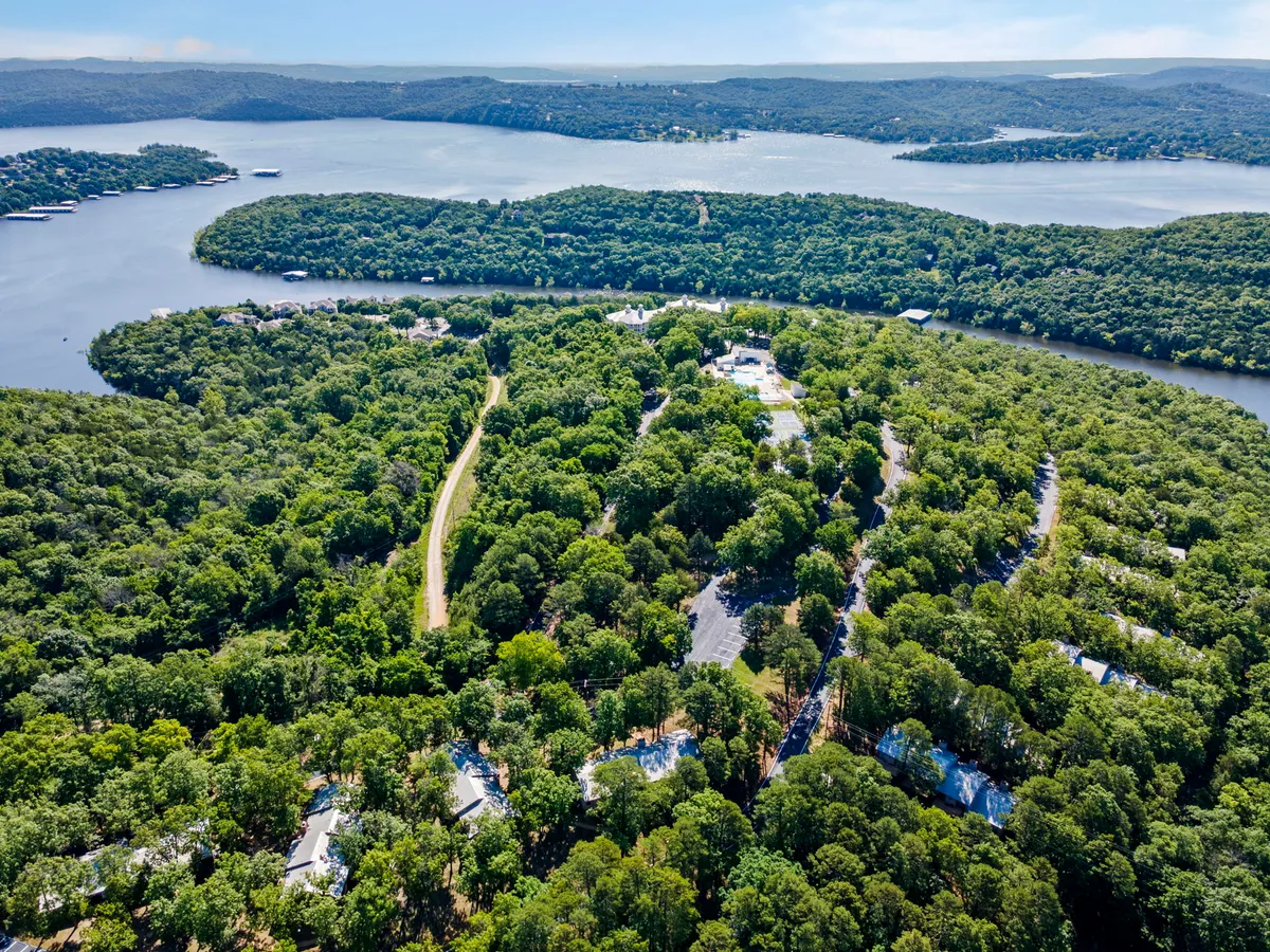 A sweeping view of lush greenery encircling a tranquil lake, with a curved road and scattered houses visible along the landscape. The water reflects the sky, and the gentle slopes suggest ample space for walking, exploring, and relaxing by the lakeside.
