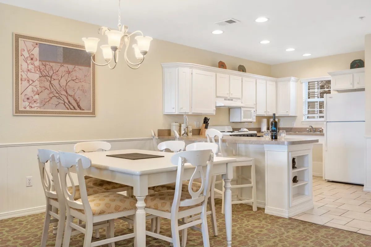 A cozy dining area with a stylish dining table, a decorative chandelier above, kitchen cabinetry in the background, and tasteful wall art. The setting feels intimate and warm, suitable for both casual dining and celebratory dinners.
